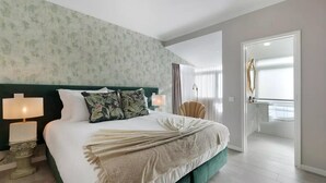 A modern hotel room with a large bed, two bedside lamps, a green headboard, floral pillows, a beige throw blanket, and a view of the bathroom through an open door. - Impeccable 4-bed House in São Domingos de Rana (Cascais)
