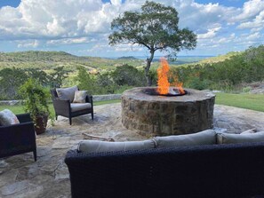 A stone fire pit with a fire, surrounded by wicker furniture and a potted plant, set on a patio with a view of a hilly landscape.
