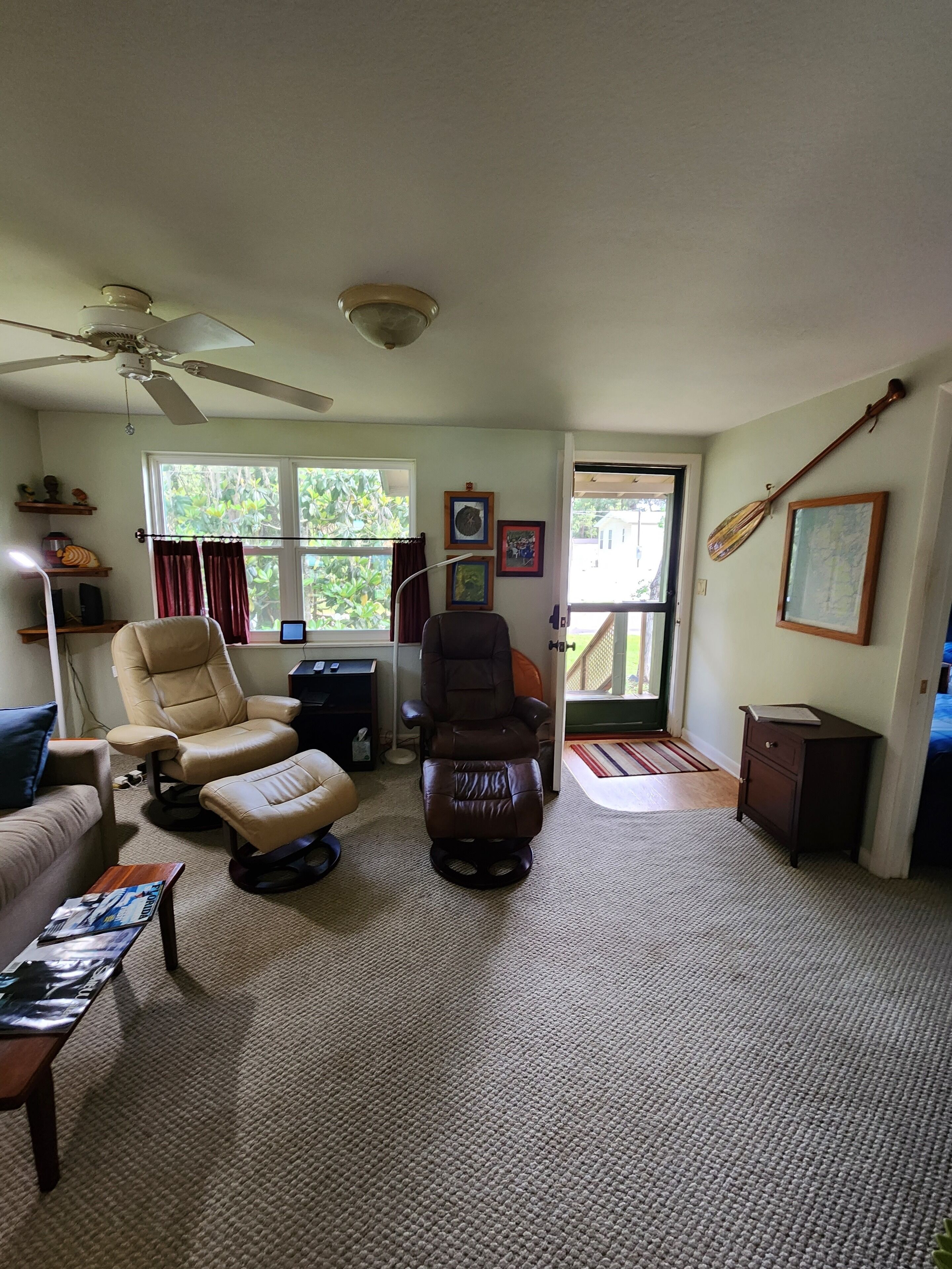 Un salon doté d’un ventilateur de plafond, de deux fauteuils en cuir, d’une petite table avec des magazines, d’un fauteuil de massage brun foncé, d’un meuble en bois et d’une grande fenêtre avec des rideaux.