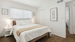 A bedroom with a bed, bedside tables, a window with blinds, and framed artwork on the walls.