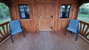 Eine Holzhütte mit einer mittleren Tür, je einem Fenster links und rechts davon, und einer Veranda mit zwei Stühlen.