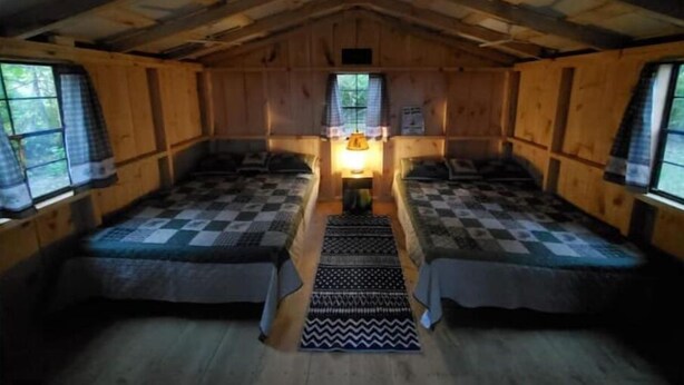 Ein gemütliches Blockhaus mit zwei Holzbett, einem zentralen Tisch mit Lampe und einem Fenster mit Vorhängen.