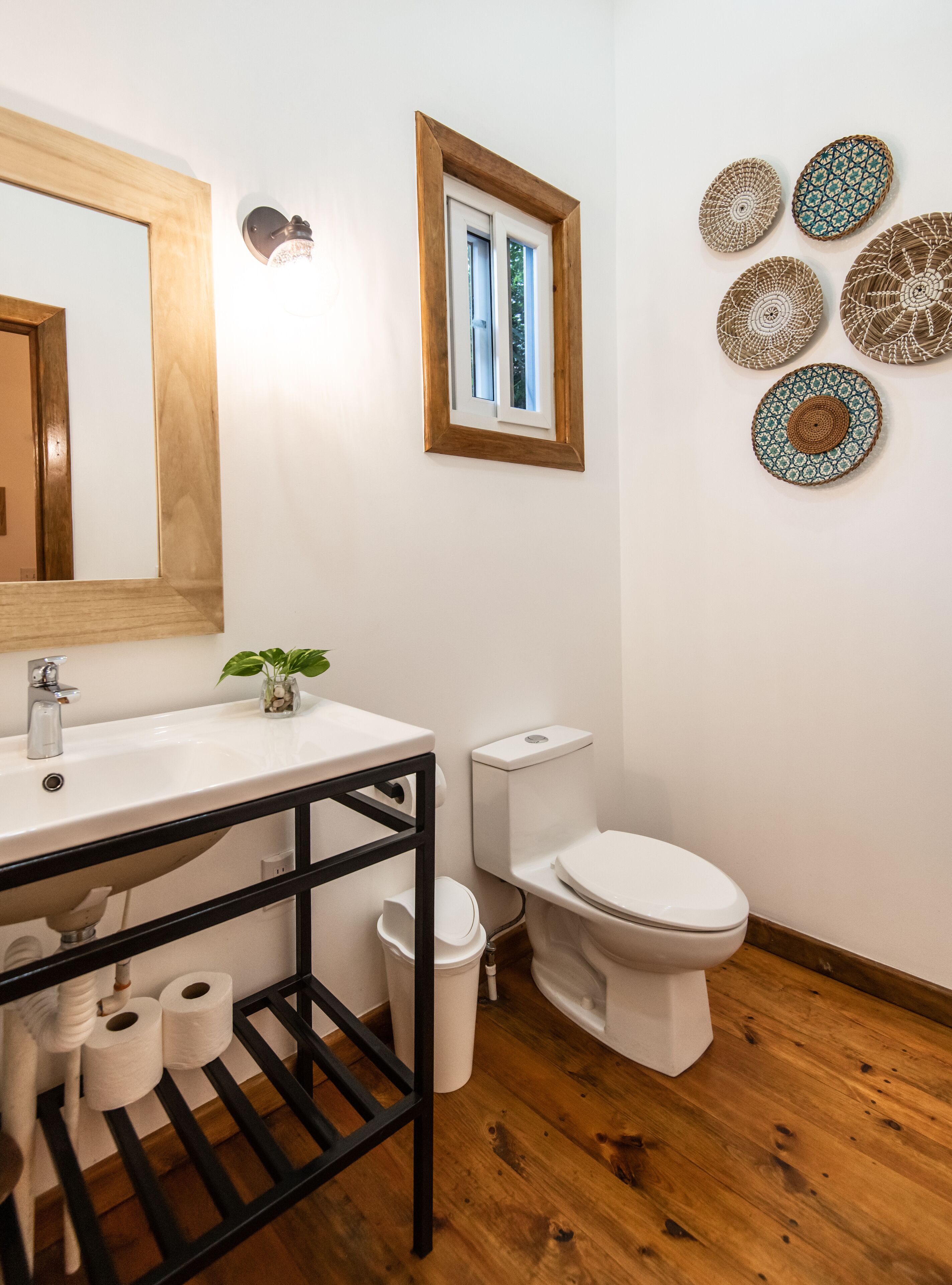 Un baño con piso de madera, paredes blancas, un espejo grande, un lavamanos con repisa negra, un inodoro y placas decorativas en la pared.