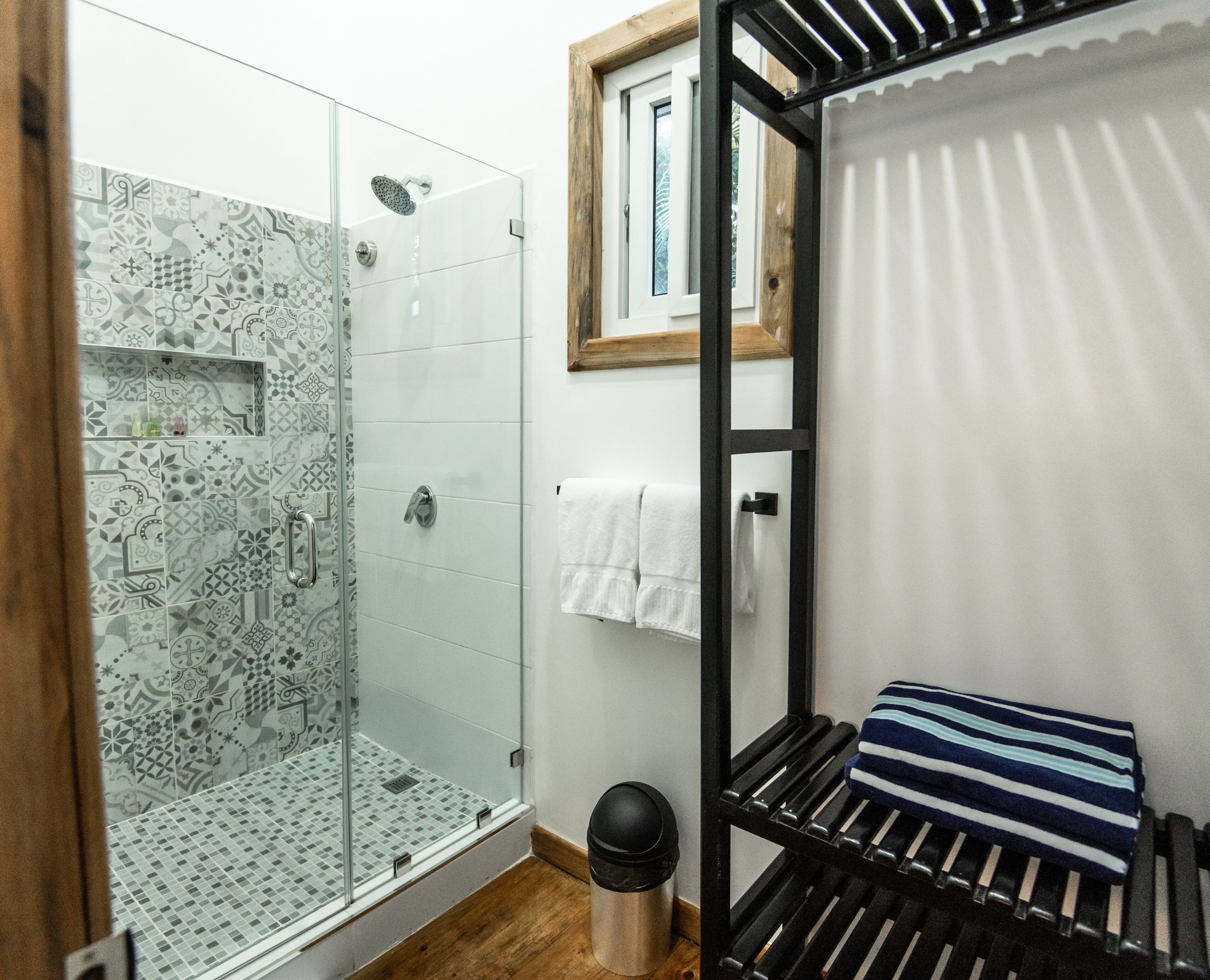 Un baño con ducha de vidrio, baldosas estampadas, un banco de madera, un perchero y un cubo de basura.
