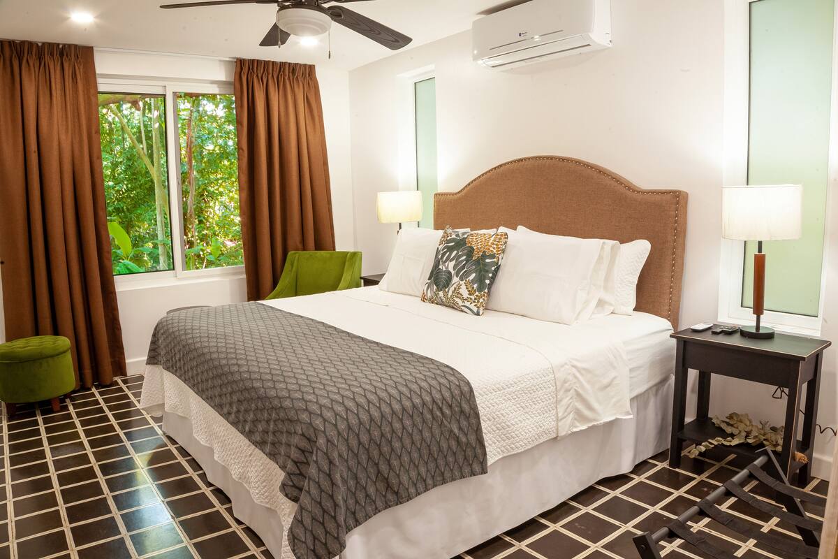 Ein Hotelzimmer mit Bett, Nachttisch, einem Fenster mit Vorhängen, Deckenventilator und Klimaanlage.