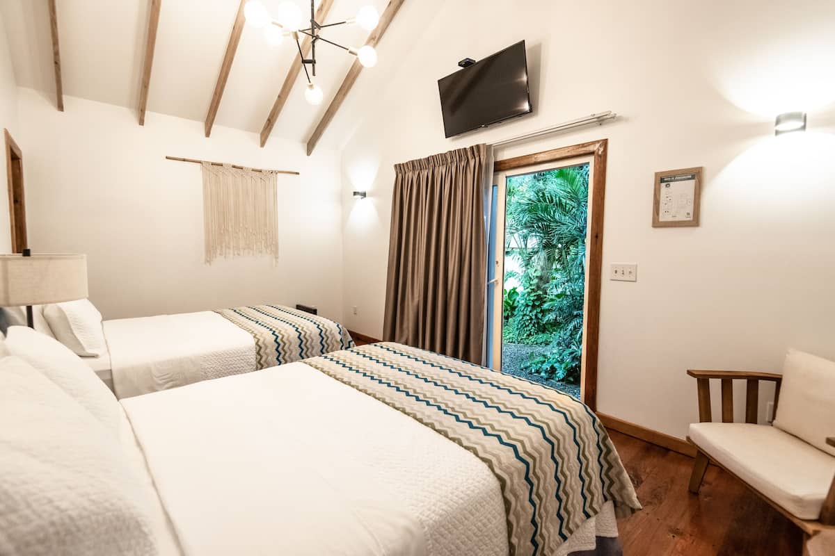 Ein Hotelzimmer mit zwei Betten, einem Fernseher, einer Holzbalkendecke, einem Fenster mit Vorhängen und Blick auf Grünflächen.