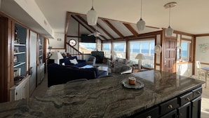 Amplia sala de estar con una isla grande, vigas de madera y vista al mar.