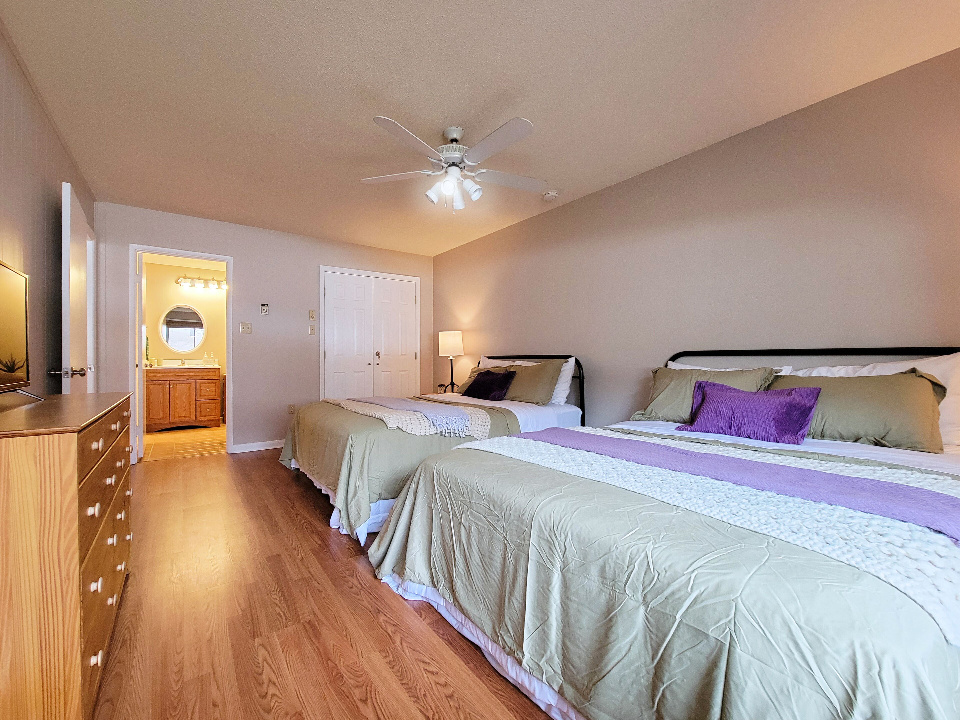 A bedroom with two beds, a ceiling fan, a bathroom with a vanity, and a wooden dresser.