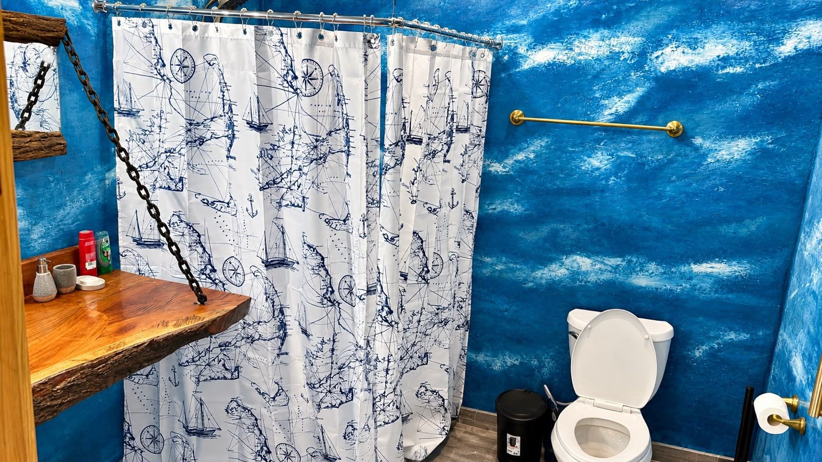 A bathroom with a nautical-themed shower curtain, a wooden shelf with toiletries, and a toilet.