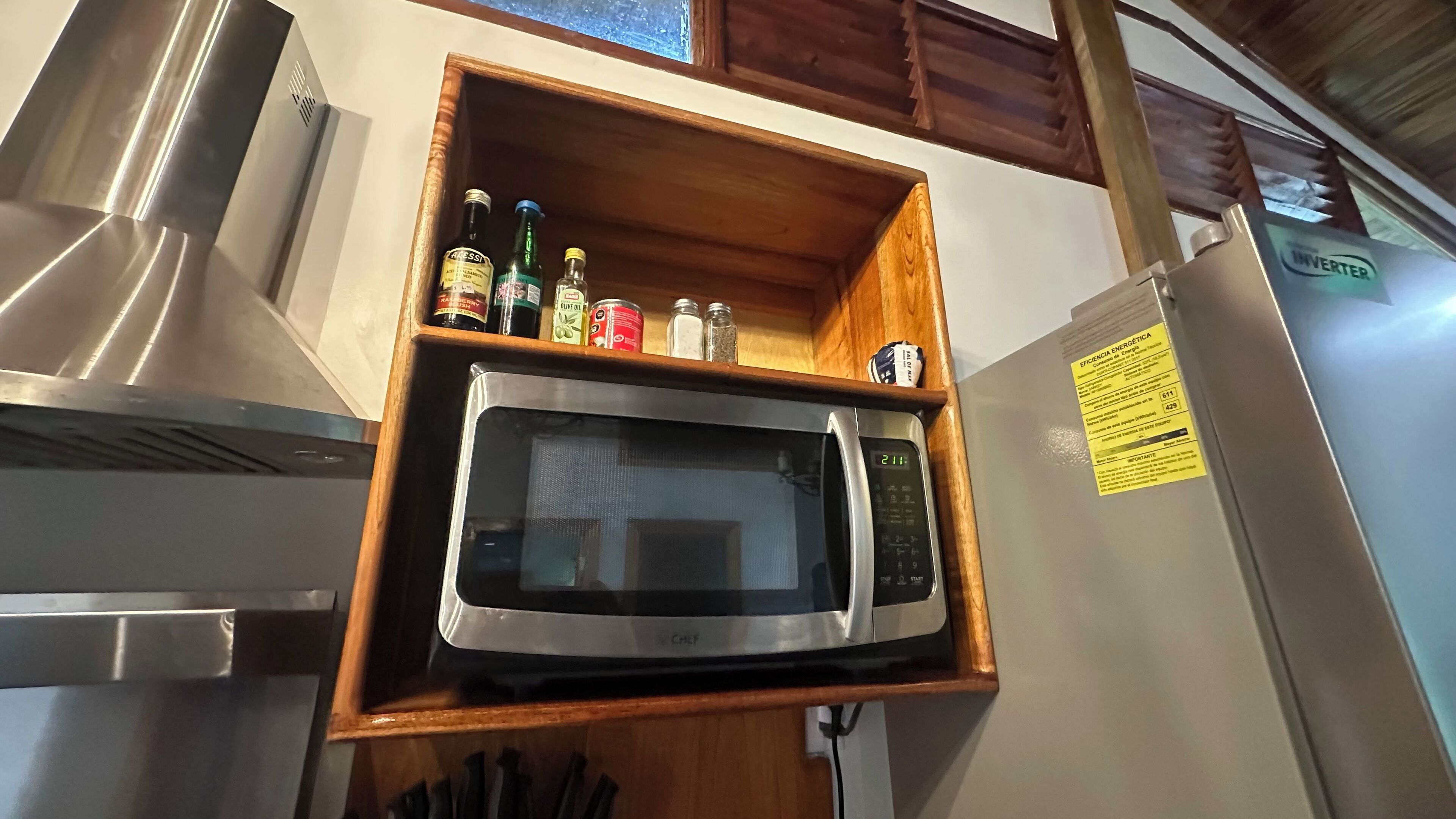 A kitchen with a built-in microwave and a wooden shelf above it containing various bottles and containers.