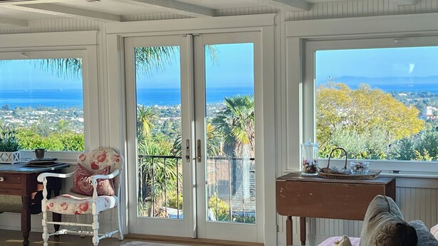 A room with a view of the ocean, a white chair, a wooden desk, and a window with a view of palm trees and houses. - OCEAN & CITY VIEWS 1920s Monthly, RIVIERA HOME, 2 QUEEN BEDS, WALK TO STATE ST (Santa Barbara)