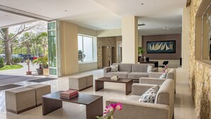 A spacious living area with a large sofa set, a coffee table, and a potted plant. There is a large window providing a view of the outside.