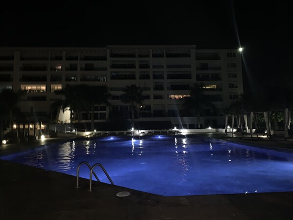 A nighttime swimming pool with illuminated water, a building with multiple floors in the background, and a few palm trees. - CASA SUNSET - Palmar’s New Luxurious 3rd Floor Seaview Condo (Cozumel)