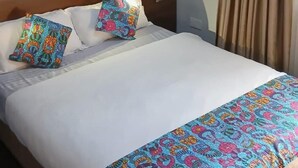Una cama bien hecha con edredón blanco y una manta decorativa de colores y diseños. Dos almohadas presentan diseños vibrantes y multicolores. La cama tiene un cabecero de madera y está ubicada frente a una pared lisa.