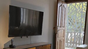 Una sala de estar con una televisión de pantalla plana montada en la pared, un mueble de madera, un tapete estampado y un balcón con una reja de metal.
