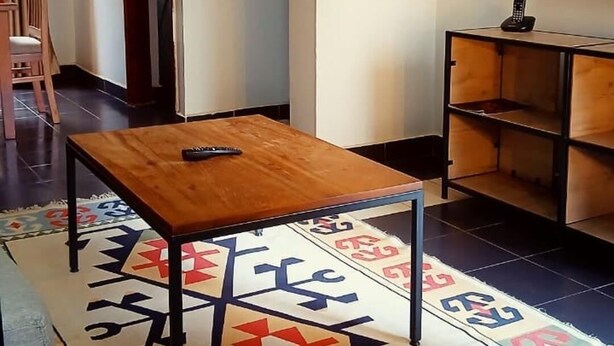 Una sala de estar con una mesa de madera, una alfombra estampada, un televisor en la pared y una estantería.