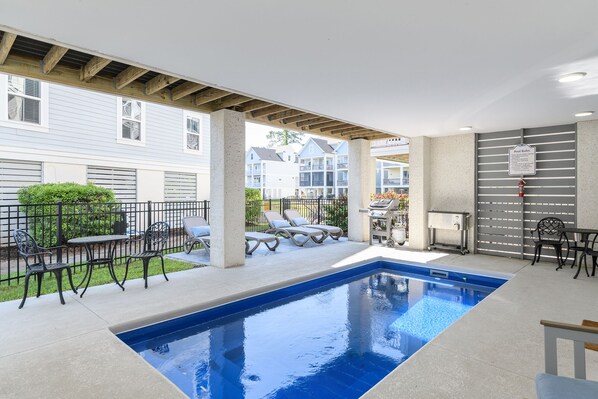 An indoor swimming pool with lounge chairs and tables, surrounded by a covered patio area.