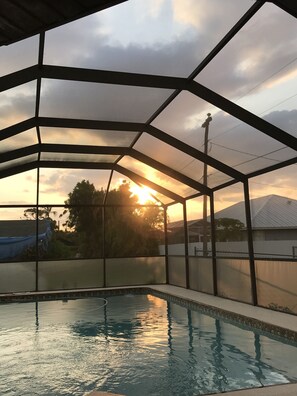 A swimming pool with a covered area, surrounded by a fence, and a sunset in the background. - VRBO House with nice solar heated pool and lanai, close to restaurants. (Cape Coral)