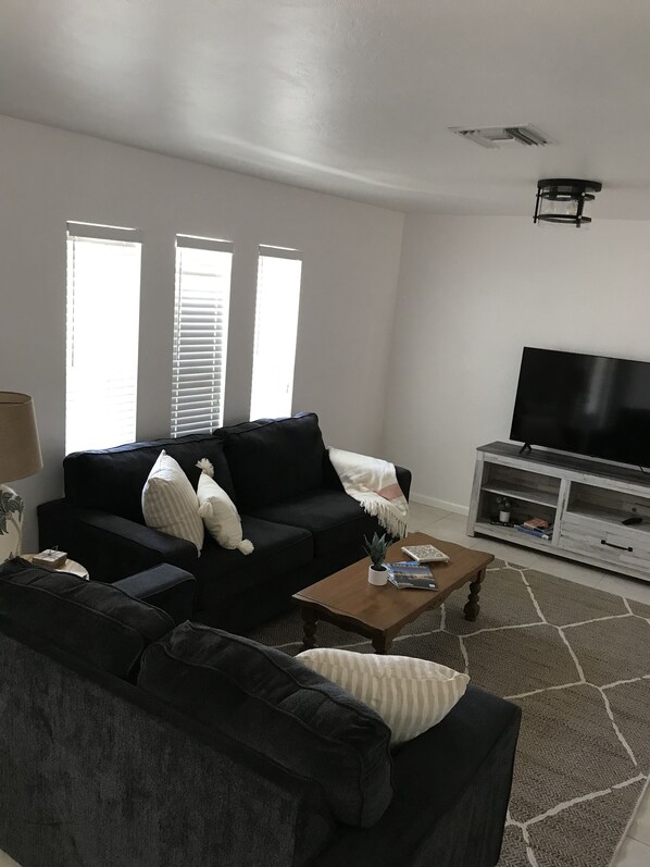 A living room with a black sofa, a wooden coffee table, a TV on a white cabinet, and a large window with blinds. - VRBO House with nice solar heated pool and lanai, close to restaurants. (Cape Coral)