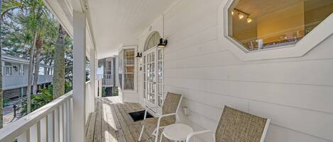 A covered porch with white wooden railings, two wicker chairs, and a view of a residential area with palm trees.