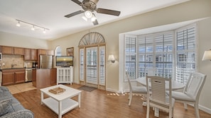 A modern kitchen with a built-in oven, a dining area with a white table and chairs, and a large window with blinds.