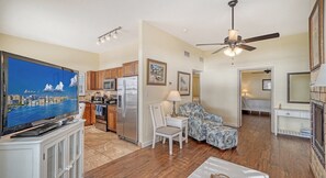 A spacious living room with a large flat-screen TV, wooden flooring, a ceiling fan, and a kitchen area with wooden cabinets and a refrigerator.