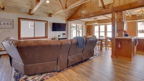 A spacious living room with a brown leather sofa, wooden ceiling beams, a flat-screen TV, and a dining area with wooden furniture. - 'Away at Moose Bay' Breezy Point Gem w/ Game Room (Breezy Point)