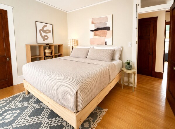 A bedroom with a wooden bed, a bedside table with a lamp, a wall shelf with decorative items, and a rug on the floor.