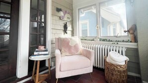 A cozy room with a pink armchair, a small side table with books, a wicker basket, and a window with a view of a house. - Rare East Side-2 min walk to UWM-Garage-Lake Mich (Milwaukee)
