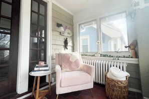 Una habitación acogedora con un sillón rosa, una mesita de noche con libros, una cesta de mimbre y una ventana con vistas a una casa.