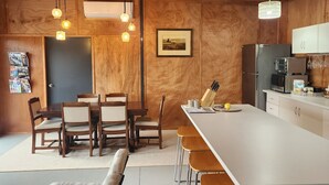 Una cocina moderna con armarios de madera, encimera blanca y taburetes de bar. Hay una zona de comedor con sillas y una mesa de madera. Un cuadro enmarcado cuelga de la pared, y hay un extintor colgado en la pared.