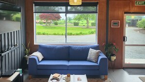 Una habitación moderna con un sofá azul, una mesita de centro con libros y una taza, un televisor a la izquierda y un amplio ventanal con vistas a zonas verdes.