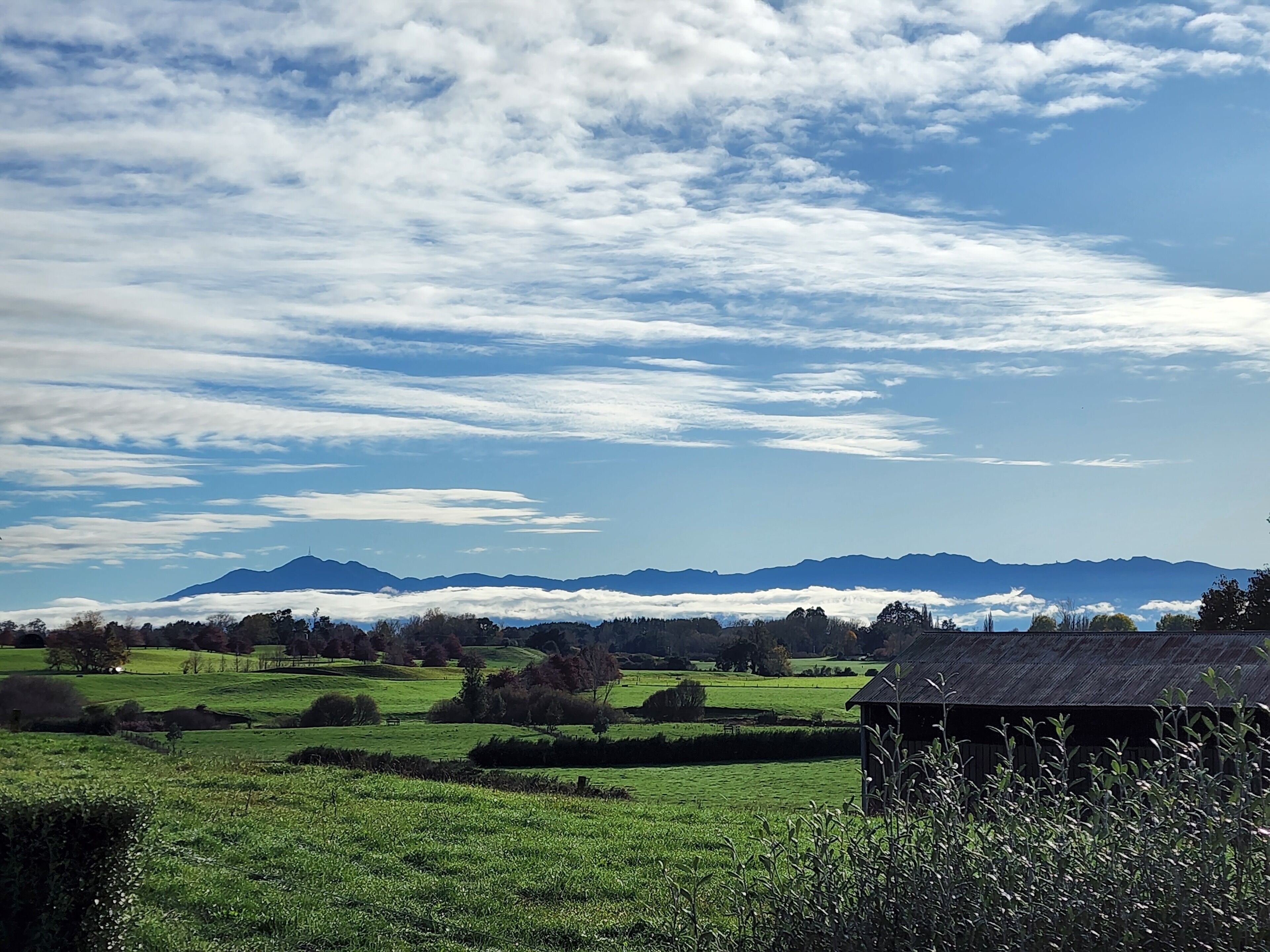 Paisagem rural com campos verdes, uma construção de madeira e montanhas ao longe sob um céu parcialmente nublado.