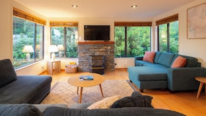 A modern living room with a stone fireplace, a flat-screen TV, and a large window offering a view of greenery. - Prime Location | 3 bedroom home | Unlimited WIFI (Queenstown)