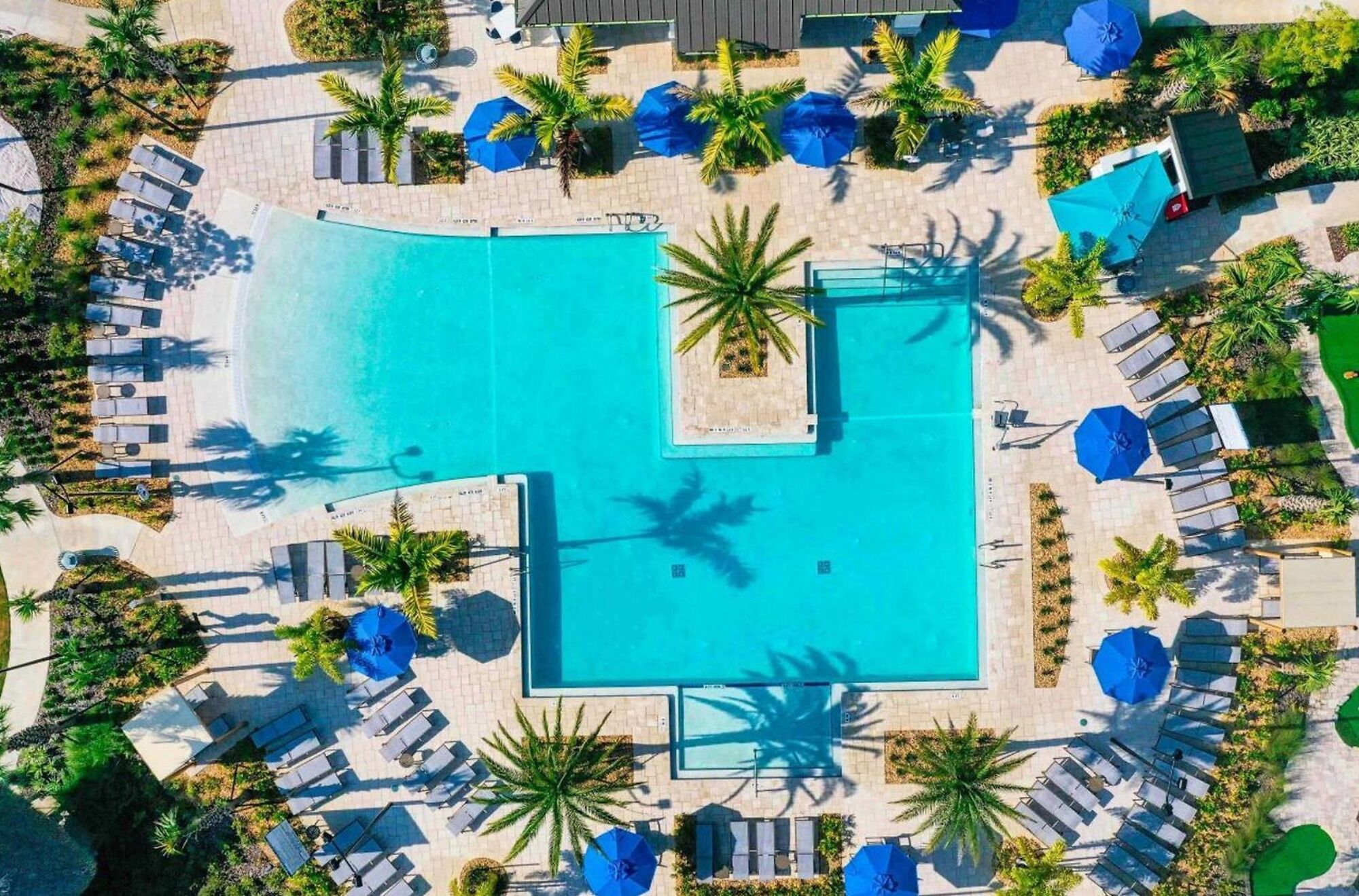 Et luftfoto af et resort med en central swimmingpool omgivet af loungeområder og palmer.