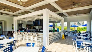 An outdoor dining area with white tables and blue chairs, a bar area with a counter, and a ceiling with wooden beams and ceiling fans. - Luxury Champions Gate Condo, Resort-Style Retreat (Four Corners)