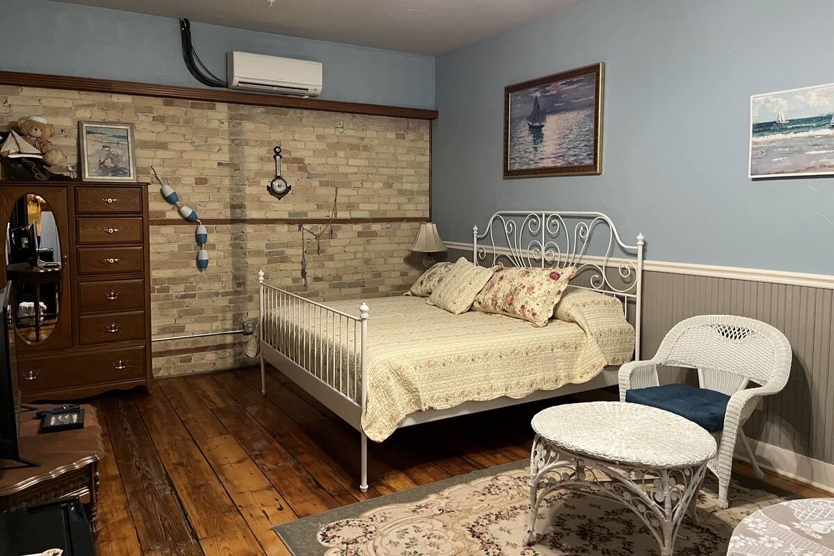 A bedroom with a brick wall, a bed with a white metal frame, a dresser, a chair, and a small round table.