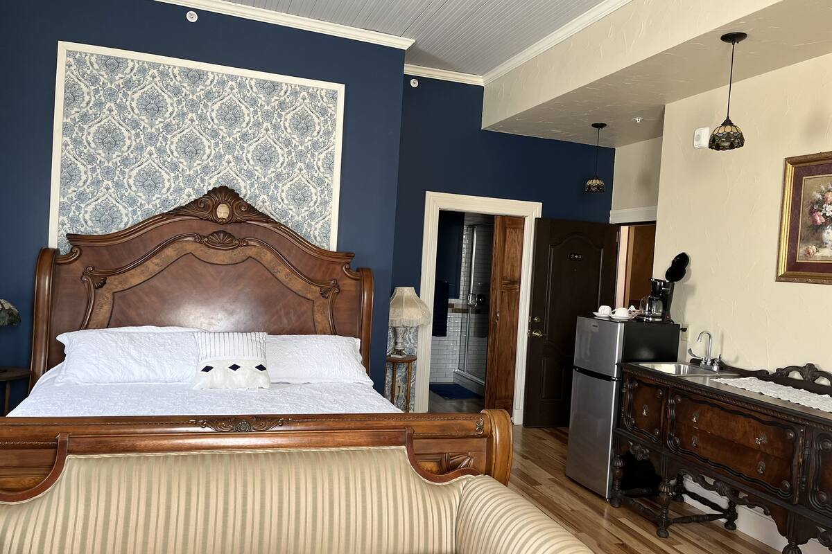 A bedroom with a large bed, a wooden dresser, a refrigerator, and a bathroom visible through an open door.