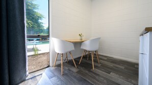 A corner room with a wooden table and two white chairs, a window with a view of a pool and trees, and a white refrigerator. - Urban Lounge Sleepery, SureStay Collection by BW (Rotorua)
