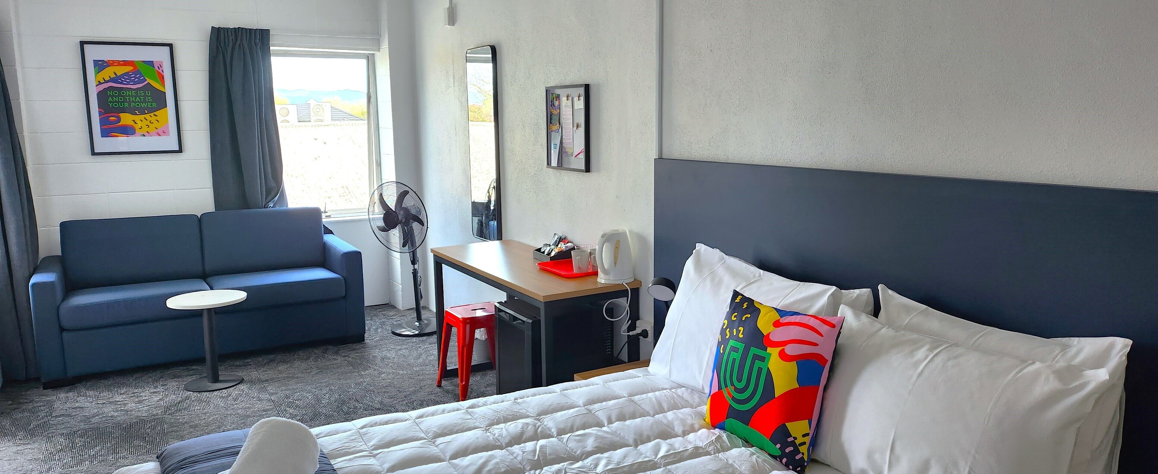 A hotel room with a bed, a desk, a chair, a blue sofa, a standing fan, and a window with curtains.
