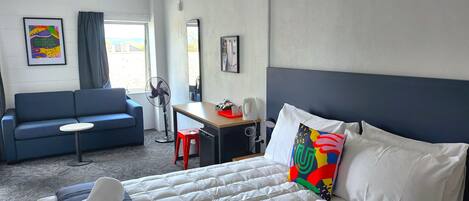 A hotel room with a bed, a desk, a chair, a blue sofa, a standing fan, and a window with curtains.