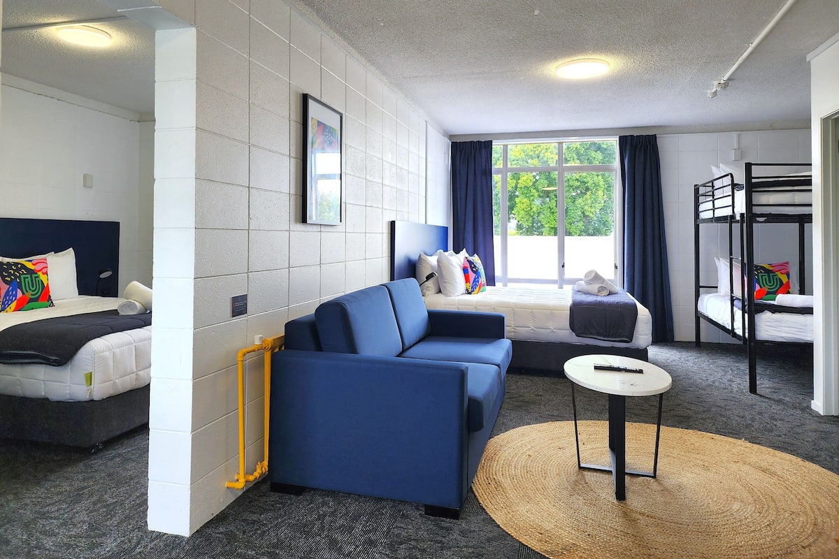 A hotel room with a sofa, a bed, a bunk bed, a small table, and a large window with curtains.