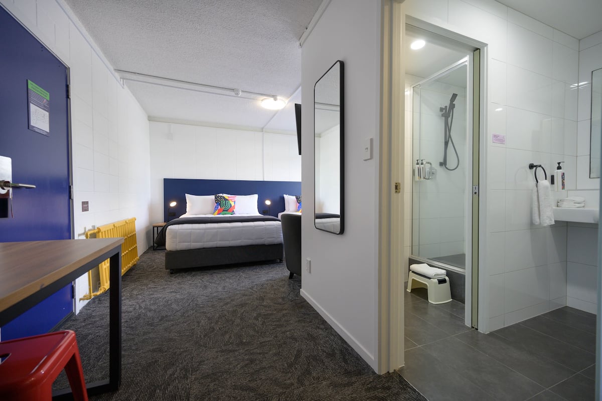 A hotel room with a bed, a shower, a small stool, and a table with a chair.