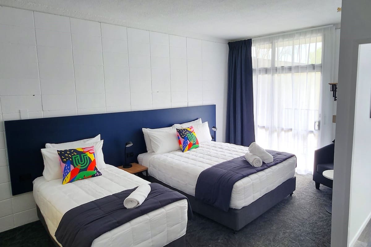 A hotel room with two beds, a standing fan, and a window with curtains.