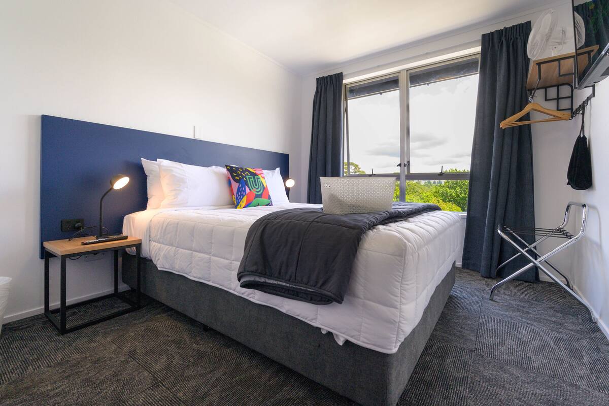 A hotel room with a large bed, a bedside table with a lamp, a window with curtains, and a wall-mounted fan.