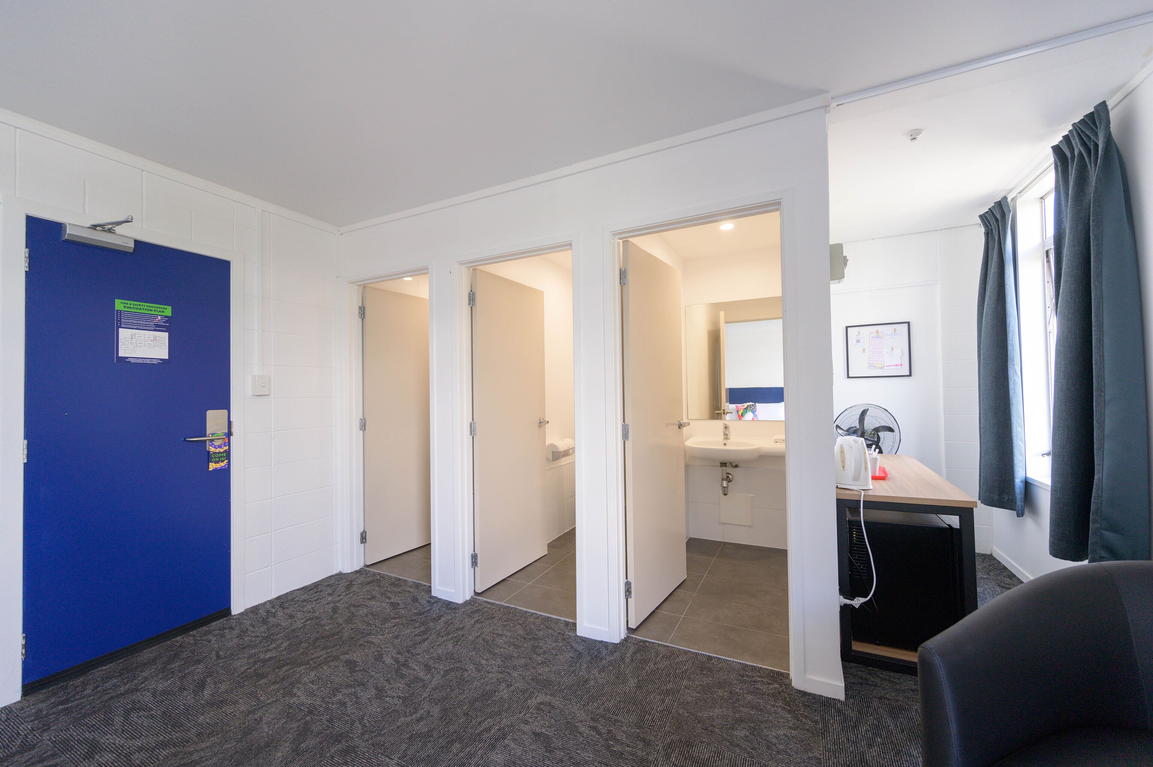 A hotel room with a blue door, a sink, a mirror, and a desk with a fan.