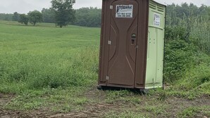 Des toilettes portables, installées sur un terrain herbeux, avec une pancarte indiquant des tarifs abordables.