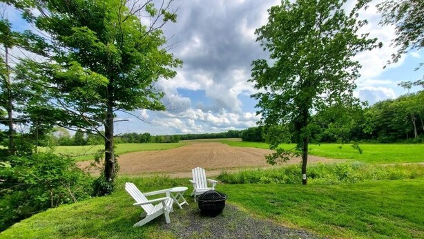 Un espace herbeux avec deux chaises Adirondack blanches et un pot noir, entouré d’arbres et d’un champ.