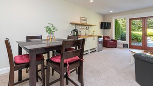 A dining area with a wooden table and chairs, a kitchen in the background, and a view of greenery through a sliding door.