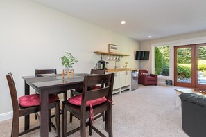 A dining area with a wooden table and chairs, a kitchen in the background, and a view of greenery through a sliding door.